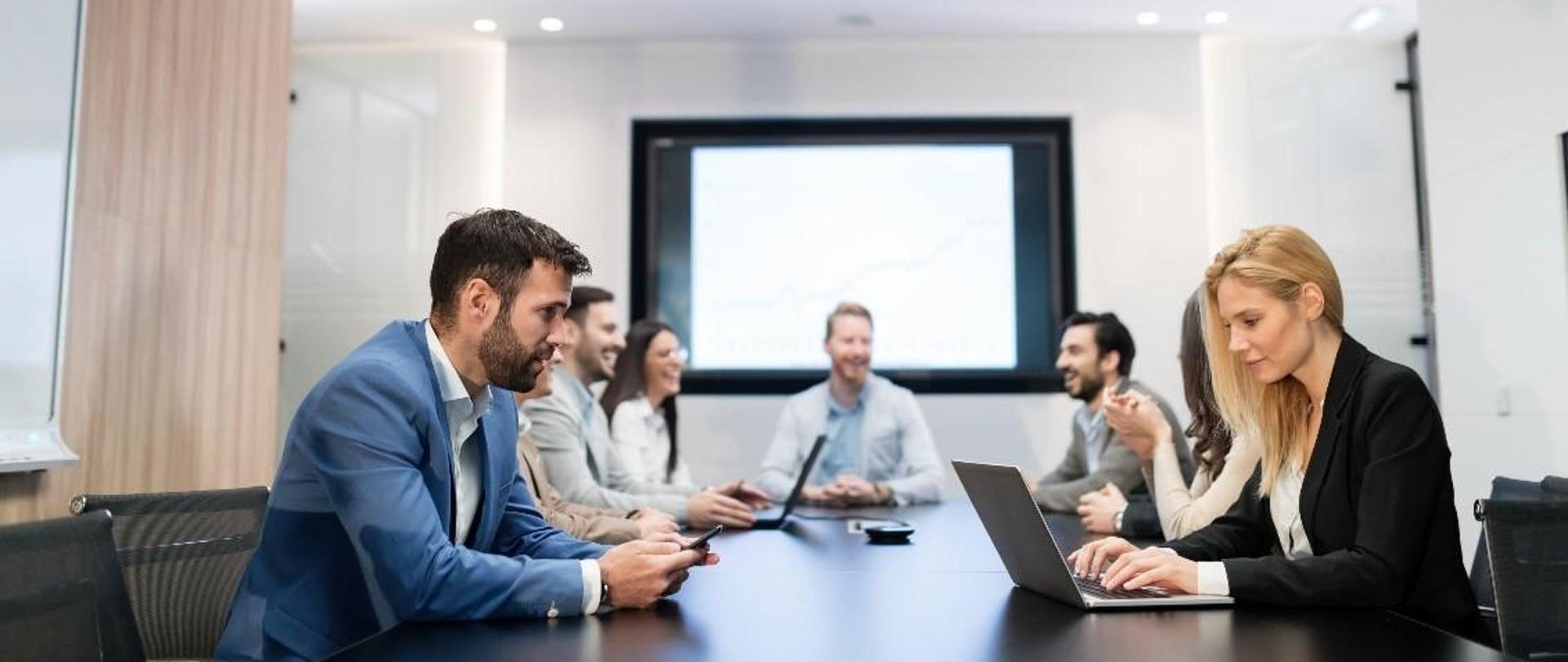 Przy stole siedzą uczestnicy spotkania, przed dwoma osobami na blacie stoją laptopy