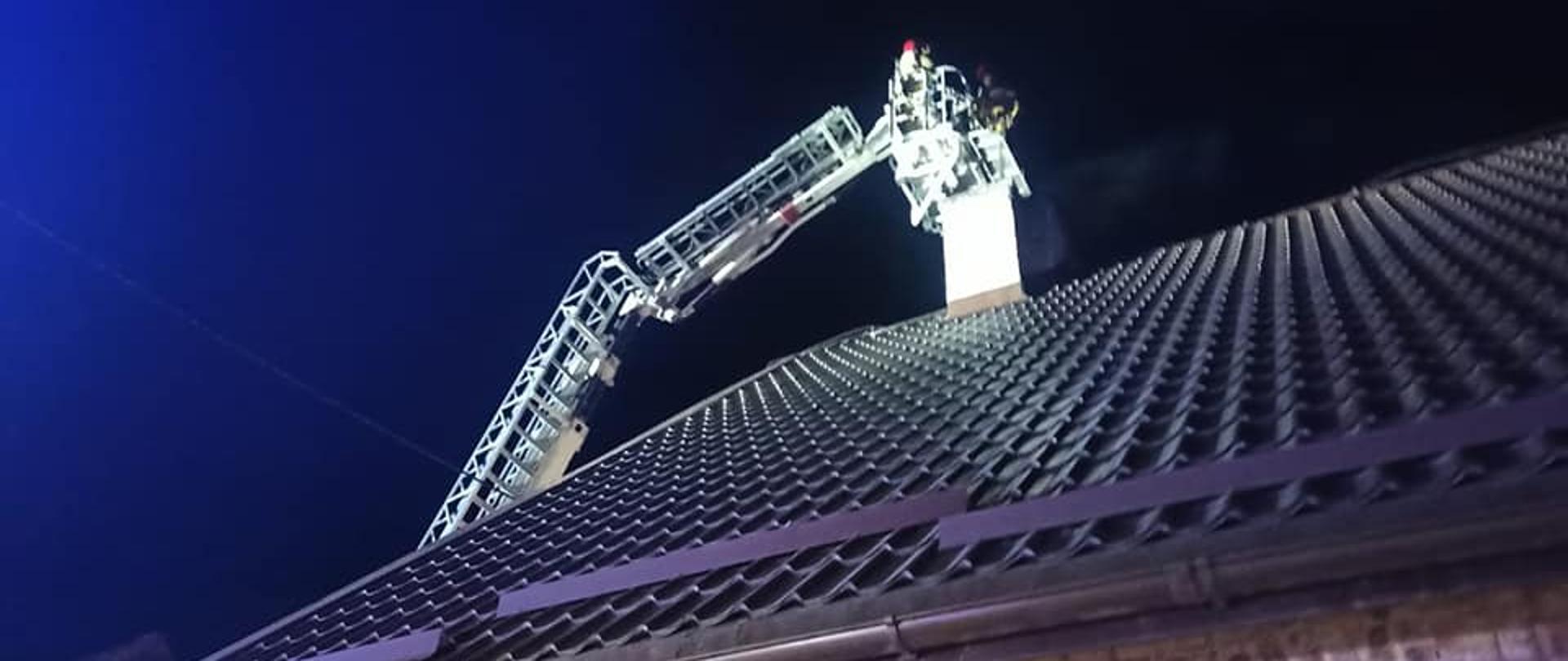 Zdjęcie wykonane w nocy. Tło stanowi ciemne niebie. Centralny punkt stanowi komin i kosz podnośnika przy kominie. W dole zdjęcia widać dach koloru brązowego. W koszu strażacy prowadzący działania gaśnicze.