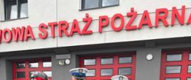 Strażak w piaskowym mundurze wręcza „Certyfikat Wsparcia” dwóm policjantom wydziału ruchu drogowego przed budynkiem Państwowej Straży Pożarnej. Obok stoi kolarz w biało-kropkowanej koszulce.