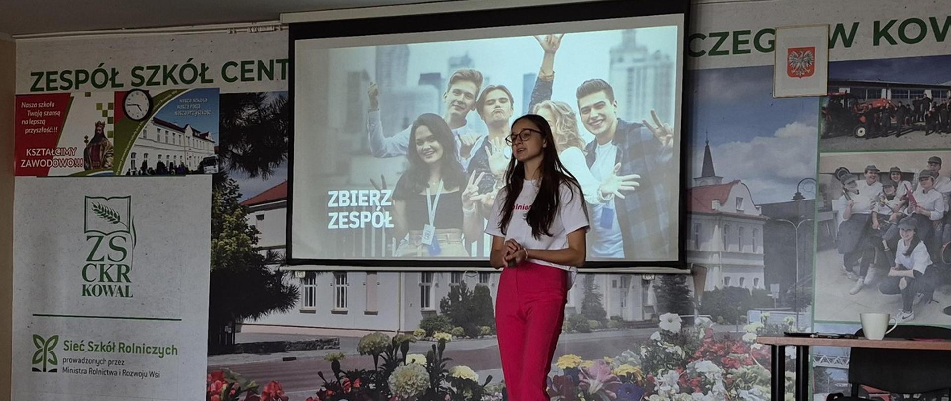 Na zdjęciu stoi kobieta ubrana w biały t-shirt oraz czerwone spodnie. Ma okulary. Przemawia do publiczności. W tle widać scianę na której widnieje napis "Zbierz zespół"