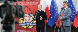 Strażak w mundurze wyjściowym stoi przy mikrofonie i przemawia obok w garniturze stoi mężczyzna trzymając teczkę za nimi stoją flagi Unii Europejskiej oraz Polski obok stoi baner Komendy Głównej Państwowej Straży Pożarnej. 