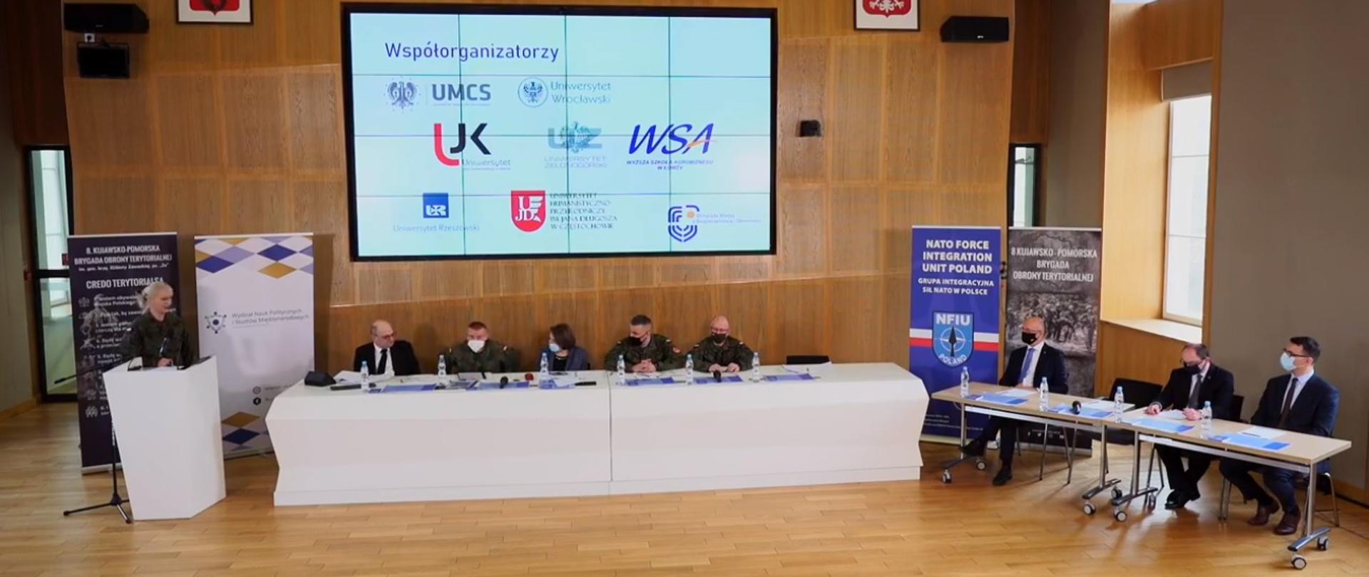 Na zdjęciu widoczna sala wykładowa wyłożona drewnianymi panelami. Od lewej mównica przy której stoi kobieta w mundurze. Na prawo od mównicy długi stół prezydialny, przy którym siedzi 5 osób (jedna kobieta, czterech mężczyzn), obok dwa stoliki, przy których siedzi trzech mężczyzn. 