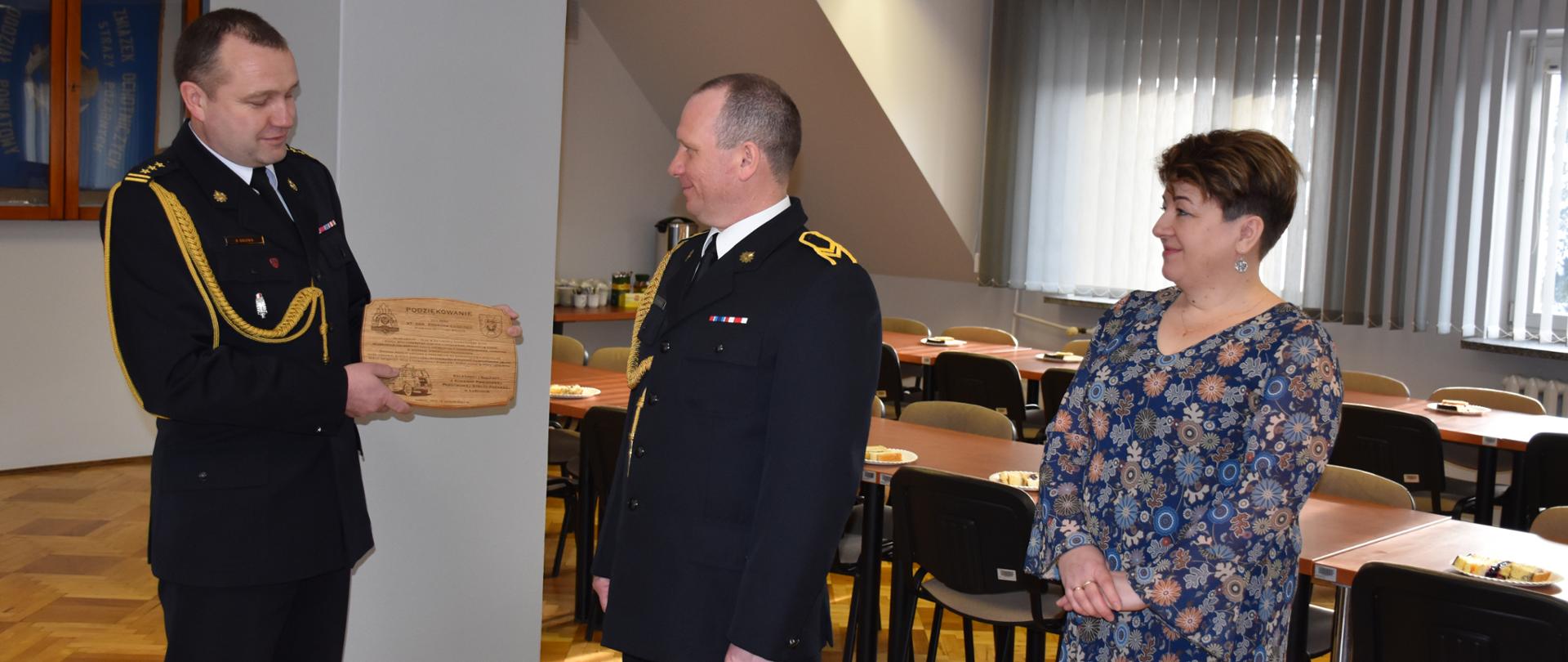 Zdjęcie przestawia pomieszczenie świetlicy Komendy Powiatowej Państwowej Straży Pożarnej w Łańcucie Na pierwszym planie od lewej: starszy brygadier Bogusław Golenia - Komendant Powiatowy Państwowej Straży Pożarnej w Łańcucie, wręczający pamiątkową deskę z podziękowaniami dla starszego ogniomistrza Konrada Cedzidło, po prawej Żona Konrada Cedzidło. W tle widoczne stoliki oraz krzesła.