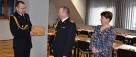 Zdjęcie przestawia pomieszczenie świetlicy Komendy Powiatowej Państwowej Straży Pożarnej w Łańcucie Na pierwszym planie od lewej: starszy brygadier Bogusław Golenia - Komendant Powiatowy Państwowej Straży Pożarnej w Łańcucie, wręczający pamiątkową deskę z podziękowaniami dla starszego ogniomistrza Konrada Cedzidło, po prawej Żona Konrada Cedzidło. W tle widoczne stoliki oraz krzesła.