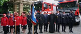 Zdjęcie przedstawia pododdziały strażaków PSP i OSP w ubraniach galowych stojące na tle trzech samochodów gaśniczych. Centralnie stoi Młodzieżowa Drużyna Pożarnicza ubrana w czerwone mundury, oraz poczet sztandarowy, następnie Ksiądz Kapelan wraz z Posłem Kazimierzem Gołojuchem i Komendantem Powiatowym PSP w Łańcucie oraz Prezesem ZOSP Adamem Reizerem. 