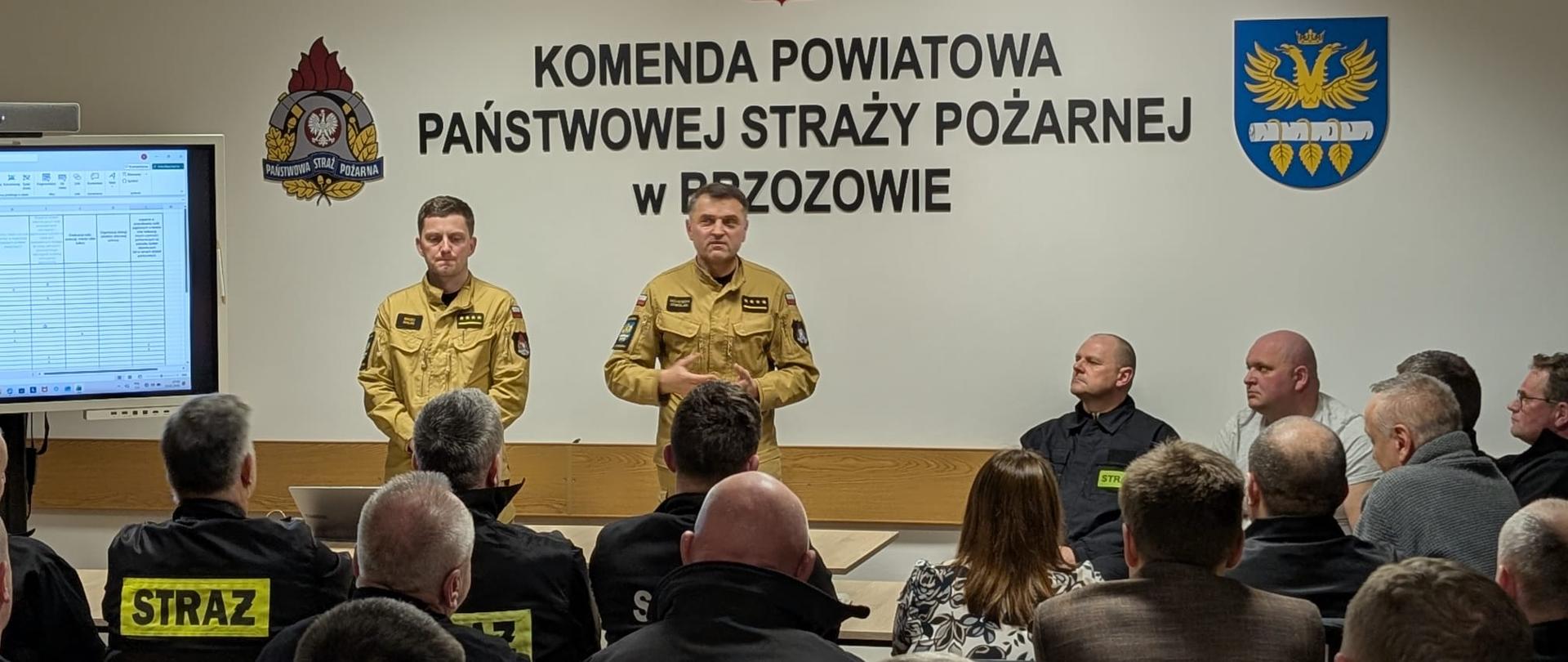 Kolorowa fotografia wykonana w pomieszczeniu świetlicy Komendy Powiatowej PSP w Brzozowie. Przedstawia dwóch strażaków PSP w strojach służbowych oraz strażaków OSP podczas narady służbowej. W tle nazwa komendy oraz logo PSP i powiatu.