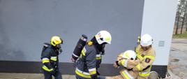 Dwóch strażaków niesie innego nieprzytomnego strażaka, który jest pozorantem w ćwiczeniu. Za nimi idzie trzeci strażak w aparacie ochrony dróg oddechowych.