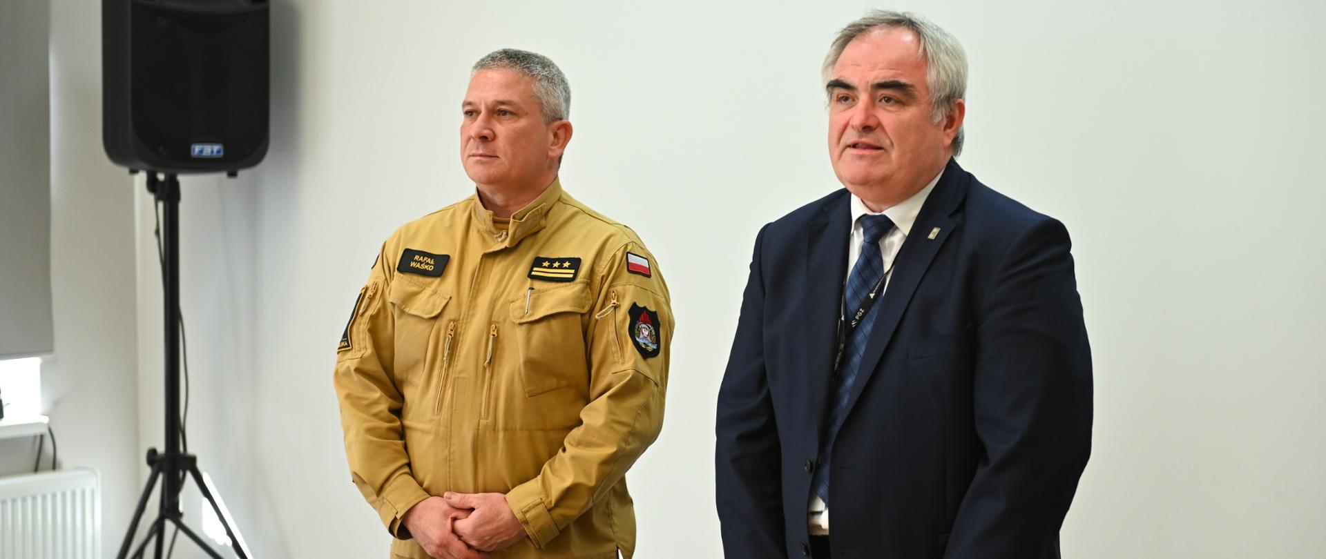 Po lewej Komendant Miejski ubrany w piaskowy mundur po prawej Prezes Zakładów Mechanicznych ubrany w ciemny garnitur. Obydwoje w postawie stojącej z tyłu ekran rzutnika 