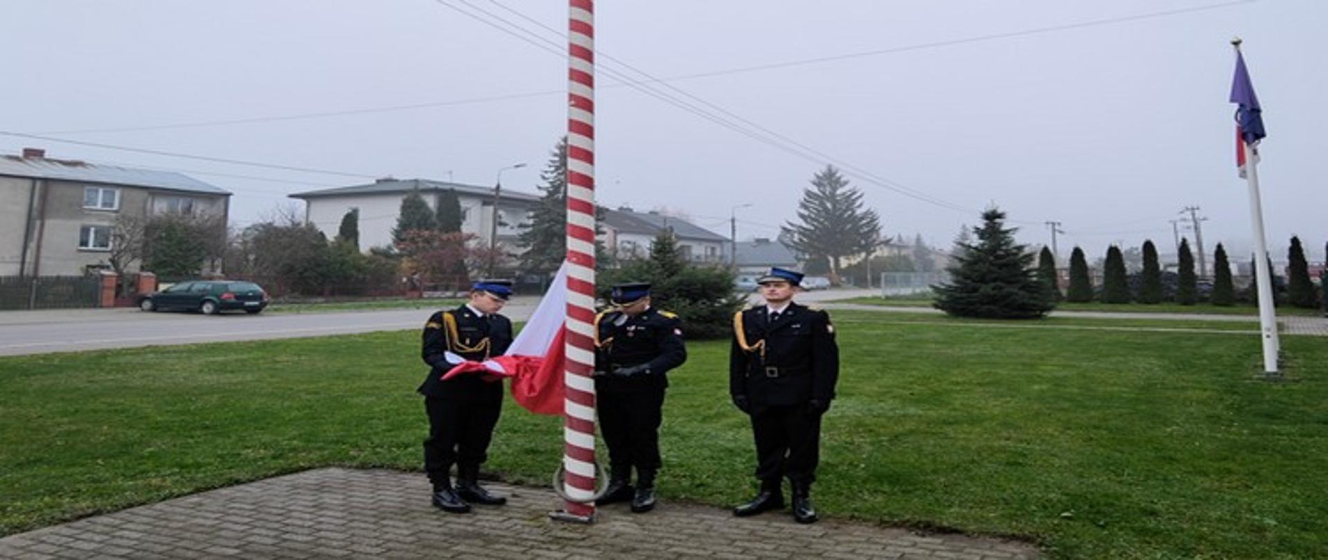 Zdjęcie przedstawia moment podnoszenia flagi państwowej na maszt w dniu 11 listopada. Poczet flagowy stanowią 3 funkcjonariusze PSP w mundurach galowych koloru czarnego. W tle zielona trawa oraz budynki mieszkalne. Pogoda pochmurna.