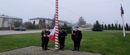 Zdjęcie przedstawia moment podnoszenia flagi państwowej na maszt w dniu 11 listopada. Poczet flagowy stanowią 3 funkcjonariusze PSP w mundurach galowych koloru czarnego. W tle zielona trawa oraz budynki mieszkalne. Pogoda pochmurna.