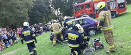 Strażacy OSP w trakcie cięcia pojazdu sprzętem hydraulicznym. Z lewej publiczność oglądająca pokaz. W tle samochód pożarniczy.