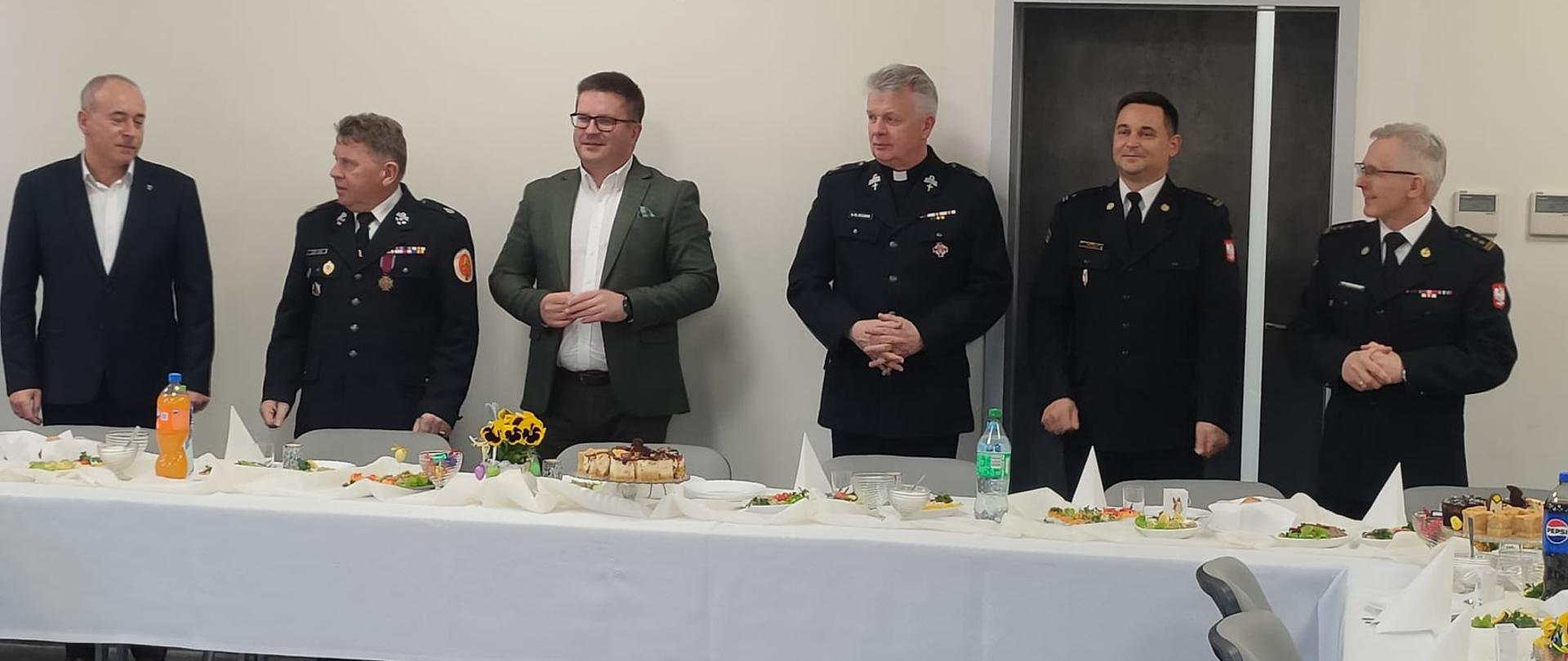 Spotkanie Wielkanocne w KP PSP w Brzezinach, które odbywa się na sali sztabowej Komendy Powiatowej Państwowej Straży Pożarnej w Brzezinach. W spotkaniu uczestniczą Komendant Powiatowy Państwowej Straży Pożarnej w Brzezinach starszy brygadier Mariusz Kaczmarski, Zastępca Komendanta Powiatowego Państwowej Straży Pożarnej w Brzezinach młodszy brygadier Jakub Brzózka, Starosta Brzeziński Bartłomiej Lipski, Burmistrz Miasta Brzeziny Dariusz Guzek, Kapelan Powiatowy Strażaków ks. Władysław Moczarski, Prezes Zarządu Oddziału Powiatowego ZOSP RP w Brzezinach dh Leszek Jarząb oraz Prezes Zarządu Koła w Brzezinach Związku Emerytów i Rencistów Pożarnictwa RP Marek, Świerczyński, wszyscy stoją przed stołami nakrytymi białymi obrusami i zastawionymi do wspólnego posiłku, na ścianie z pozującymi wisi godło Polski.