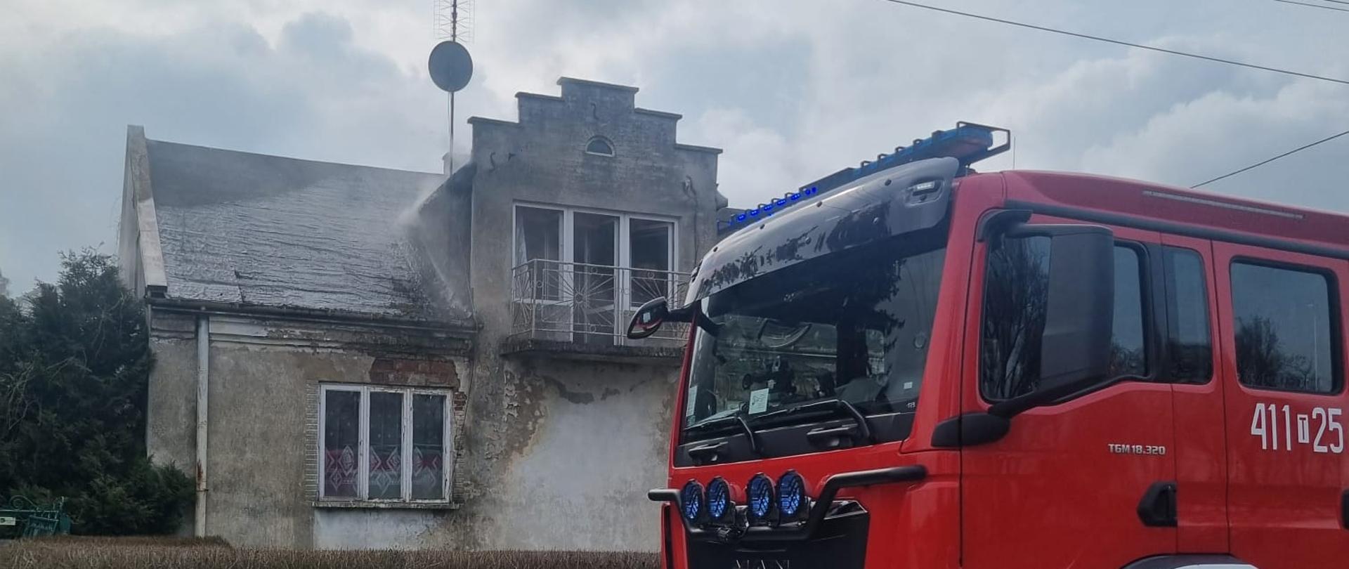 Budynek parterowy, jednorodzinny, którego dach znajduje się w lekkim zadymieniu. Przed posesją zaparkowany samochód ratowniczo-gaśniczy.
