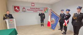 Strażak podczas ślubowania stoi z podniesioną prawą ręką. Komendant przy mównicy odczytuje rotę ślubowania. Poczet sztandarowy stoi ze sztandarem