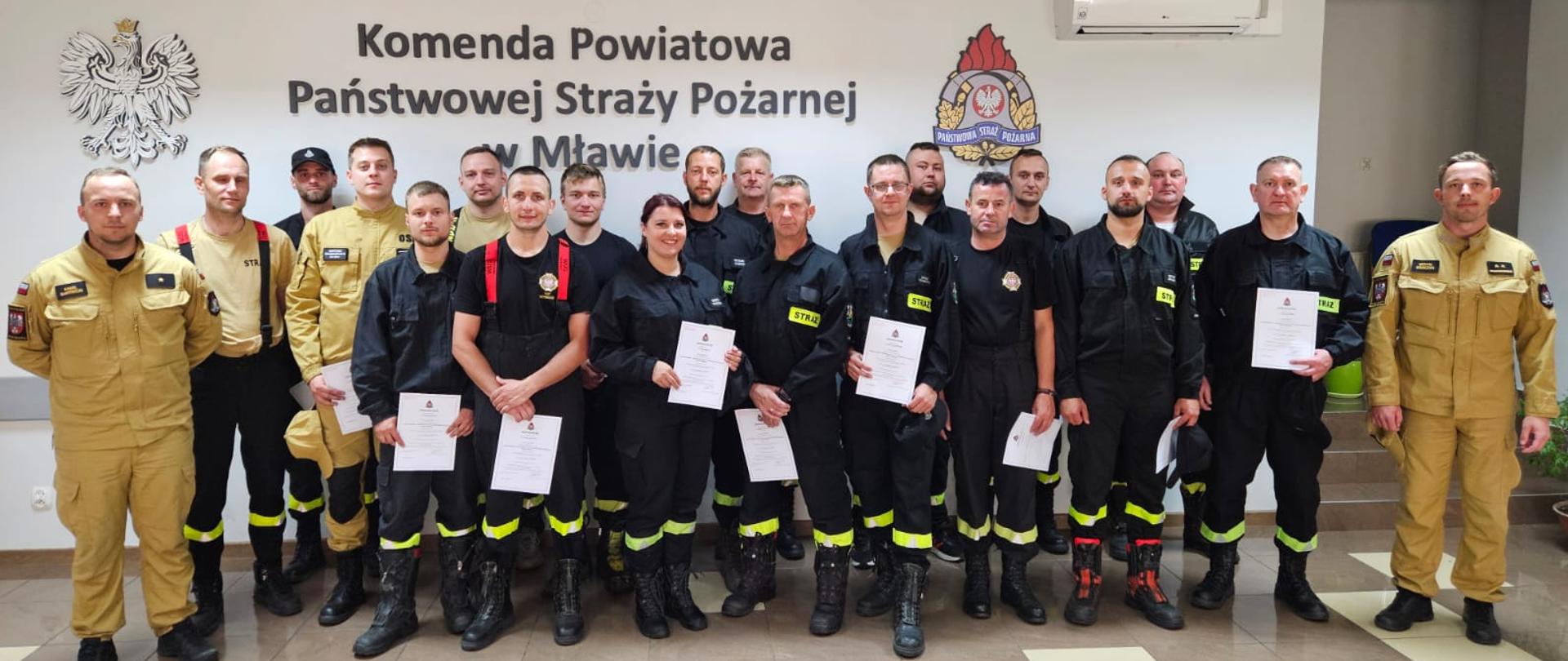 Duża grupa strażaków pozuje do zdjęcia w pomieszczeniu, stojąc w trzech rzędach. Trzech mężczyzn po lewej stronie ubranych jest w piaskowe mundury strażackie, jeden z nich nosi piaskową koszulkę polo. Reszta ubrana jest w ciemne mundury strażackie, z wyjątkiem jednej kobiety, która również nosi ciemny mundur. Prawie wszyscy trzymają w rękach białe kartki papieru, prawdopodobnie certyfikaty lub dyplomy. W tle widnieje ściana z napisem „Komenda Powiatowa Państwowej Straży Pożarnej w Mławie” oraz loga.