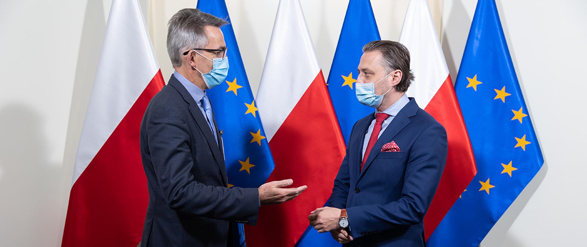 Na zdjęciu widać wiceministra Bartosza Grodeckiego i Oliviera Onidim zastępcę dyrektora generalnego DG HOME stojących i rozmawiających na tle flag Polski i UE.