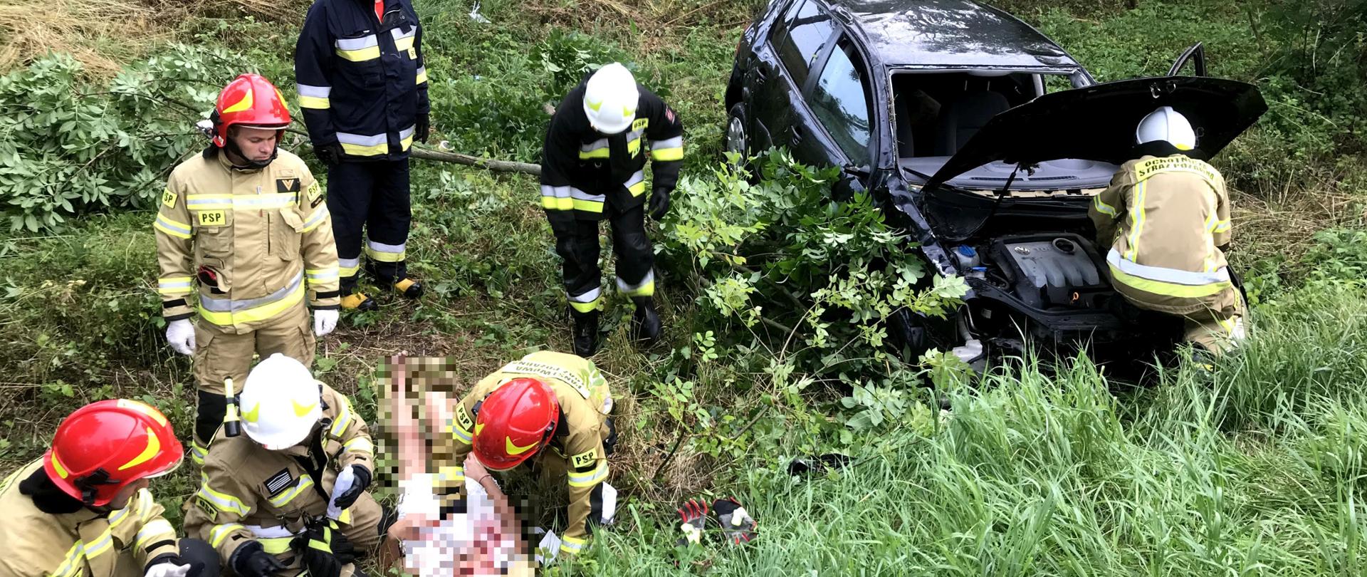 Czarny samochód osobowy znajduje się poza droga na terenie zielonym pomiędzy drzewami. Trzech strażaków udziela pomocy poszkodowanej osobie, która leży na trawie pozostali strażacy zajmują się usuwaniem kawałka złamanego drzewa oraz otwierają maskę samochodu 