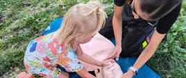 Kilkuletnia dziewczynka wraz z strażaczką uczy się pierwszej pomocy na fantomie, razem odchylają głowę. Dziewczynka o jasnych włosach i w kwiaciastej sukience. Fantom leży na trawie. 