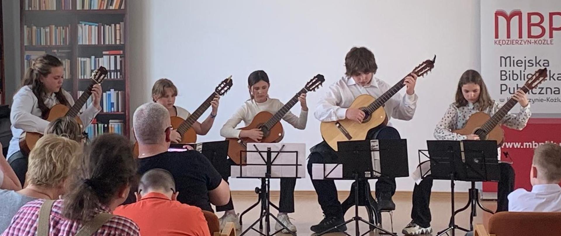 Na zdjęciu widać scenę, na której występuje pięcioro młodych ludzi grających na gitarach klasycznych. Wszyscy są skupieni na swoich instrumentach i nutach.
W tle, po lewej stronie, widać fototapetę przedstawiającą półki z książkami. Po prawej stronie wisi biało-czerwony baner Miejskiej Biblioteki Publicznej w Kędzierzynie-Koźlu.
Na pierwszym planie i w środkowej części zdjęcia siedzą słuchacze, dorośli i dzieci, którzy są odwróceni plecami do widza.