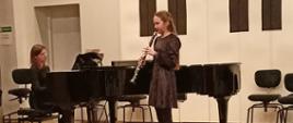 Dziewczynka stojąc na estradzie auli PSM gra na klarnecie, za nią na fortepianie gra kobieta.