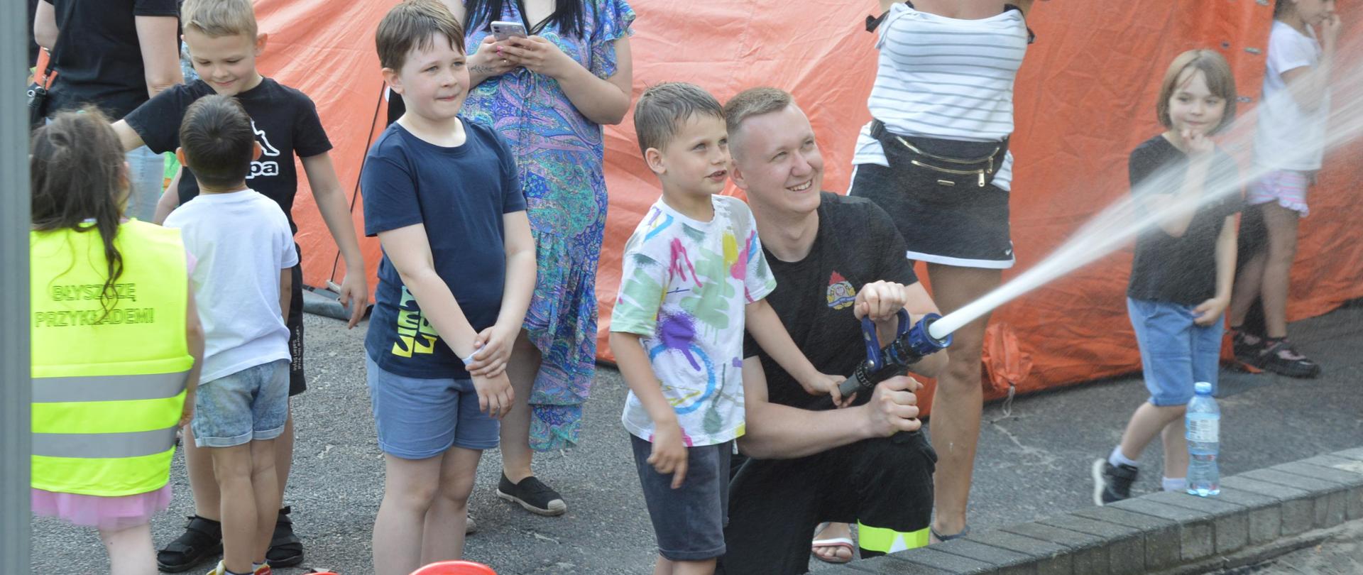 Plac wewnętrzny Komendy Powiatowej PSP w Rawiczu, na którym ustawiony jest pomarańczowy namiot pneumatyczny. Obok niego znajduje się "stanowisko gaśnicze", które obsługują dzieci uczestniczące w dniu otwartym. Mama jednego z "prądowników" wykonuje zdjęcie. Na murku leżą dwa hełmy.