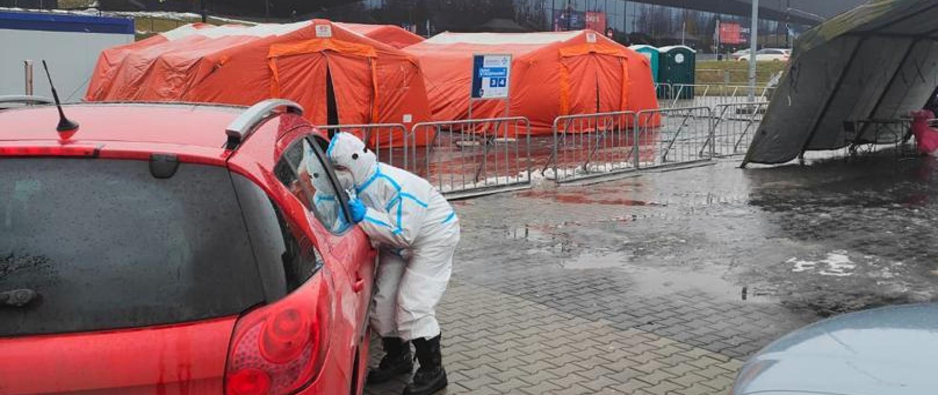 Strażak w ubraniu ochronnym pochyla się przy samochodzie w miejscu gdzie uruchomiono punkt wymazowy, w tym drive thru