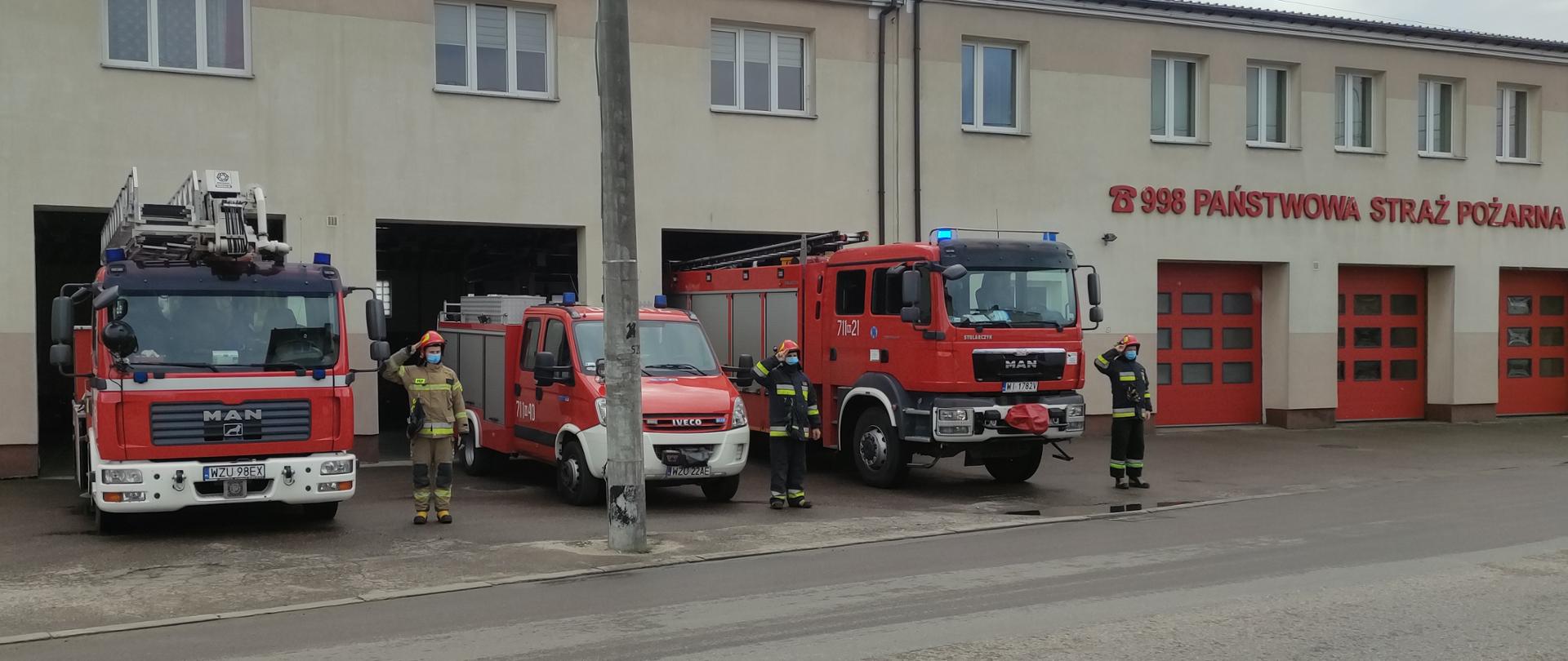 Strażacy upamiętnili Policjanta zastrzelonego na służbie stojąc przed garażami samochodami ratowniczo-gaśniczymi z włączonymi sygnałami świetlnymi i dźwiękowymi. Trzech strażaków w strojach specjalnym stoi przy samochodach salutując. 