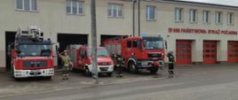 Strażacy upamiętnili Policjanta zastrzelonego na służbie stojąc przed garażami samochodami ratowniczo-gaśniczymi z włączonymi sygnałami świetlnymi i dźwiękowymi. Trzech strażaków w strojach specjalnym stoi przy samochodach salutując. 