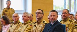 Mężczyzna w granatowym garniturze siedzi w pierwszym rzędzie obok grupy funkcjonariuszy Państwowej Straży Pożarnej w mundurach służbowych. Uczestnicy wydarzenia są zwróceni w stronę prowadzącego.