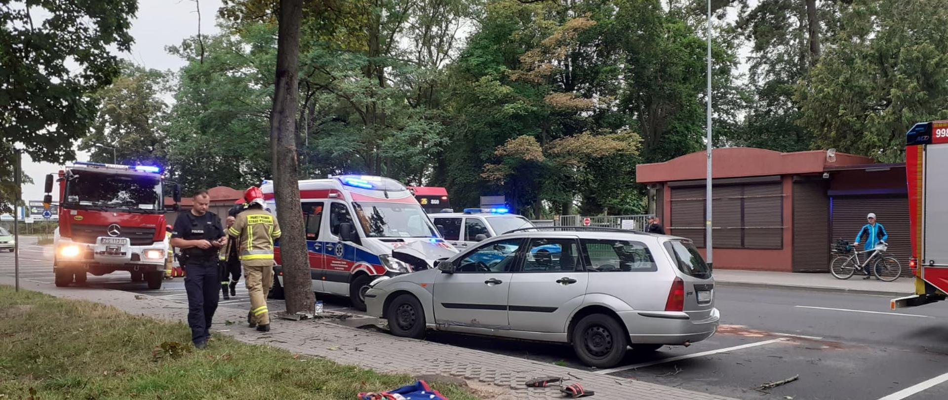 Rozbity samochód przed drzewem, widać również samochód pożarniczy karetkę pogotowia oraz koguty radiowozu.