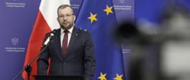 Minister funduszy i polityki regionalnej Grzegorz Puda przemawia do mikrofonu, za jego plecami flagi Polski i UE oraz baner MFiPR