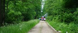 Widoczne tereny leśne, drogą leśna idzie linia gaśnicza, w oddali widoczny pojazd gaśniczy podający wodę oraz strażacy i służby leśne. 