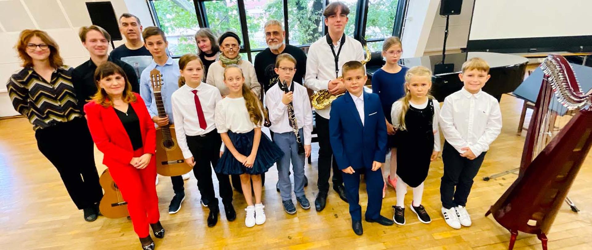 Na zdjęciu grupa kilkunastu osób, dorosłych i dzieci, stojących w sali z podłogą z parkietu. Za nimi ściana z dużym oknem, przy oknie z prawej strony stoi fortepian, z prawej strony osób stoi harfa. Kilkoro dzieci na zdjęciu trzyma w rękach instrumenty muzyczne - gitary, klarnet i saksofon.