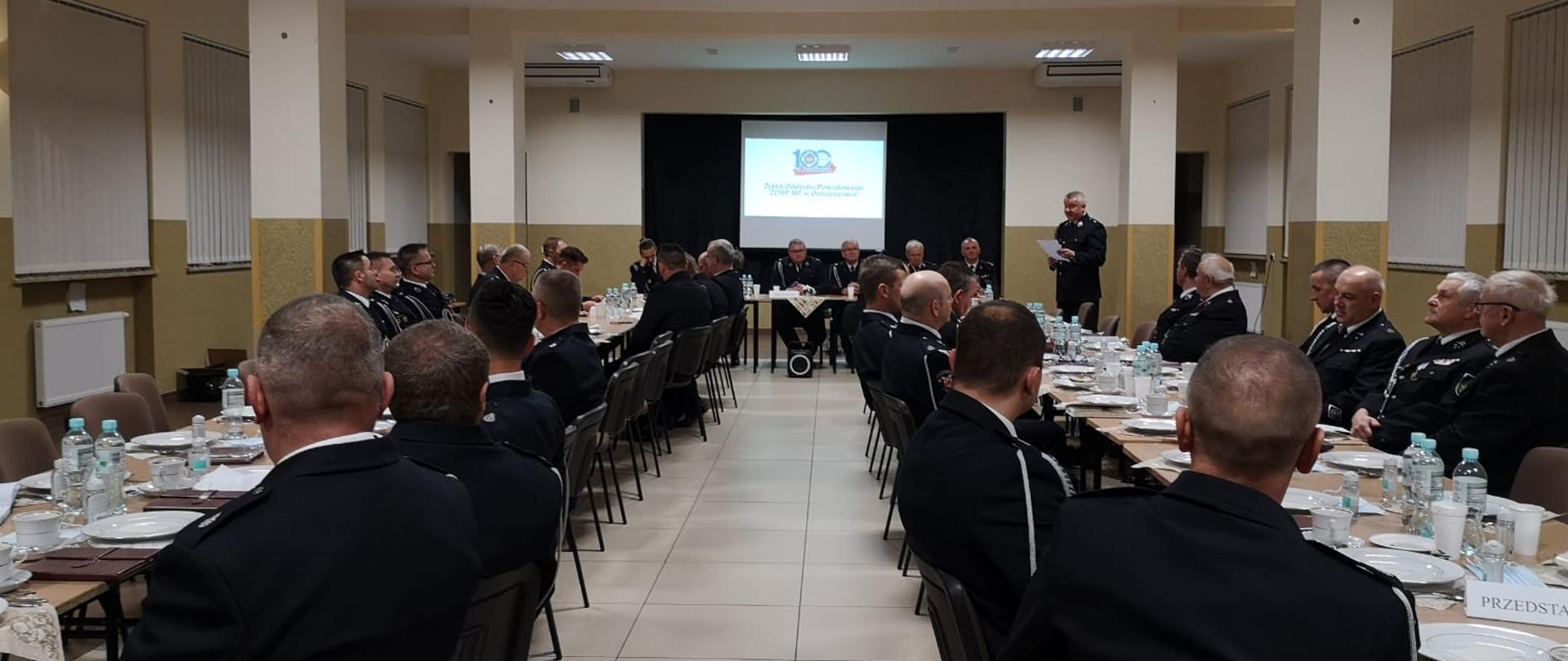 Na zdjęciu druhowie siedzący tyłem do fotografa na sali, w trakcie zjazdu oddziału powiatowego. Osoby ubrane w mundury galowe. Na dalszym planie wyświetlony slajd na ekranie projektora.