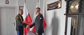 Pomorski komendant wojewódzki Państwowej Straży Pożarnej ściska dłoń strażakowi w drugiej ręce trzyma teczkę za nimi stoją dwie flagi Polski na ścianie wisi szabla oraz niedaleko stoi zegar.