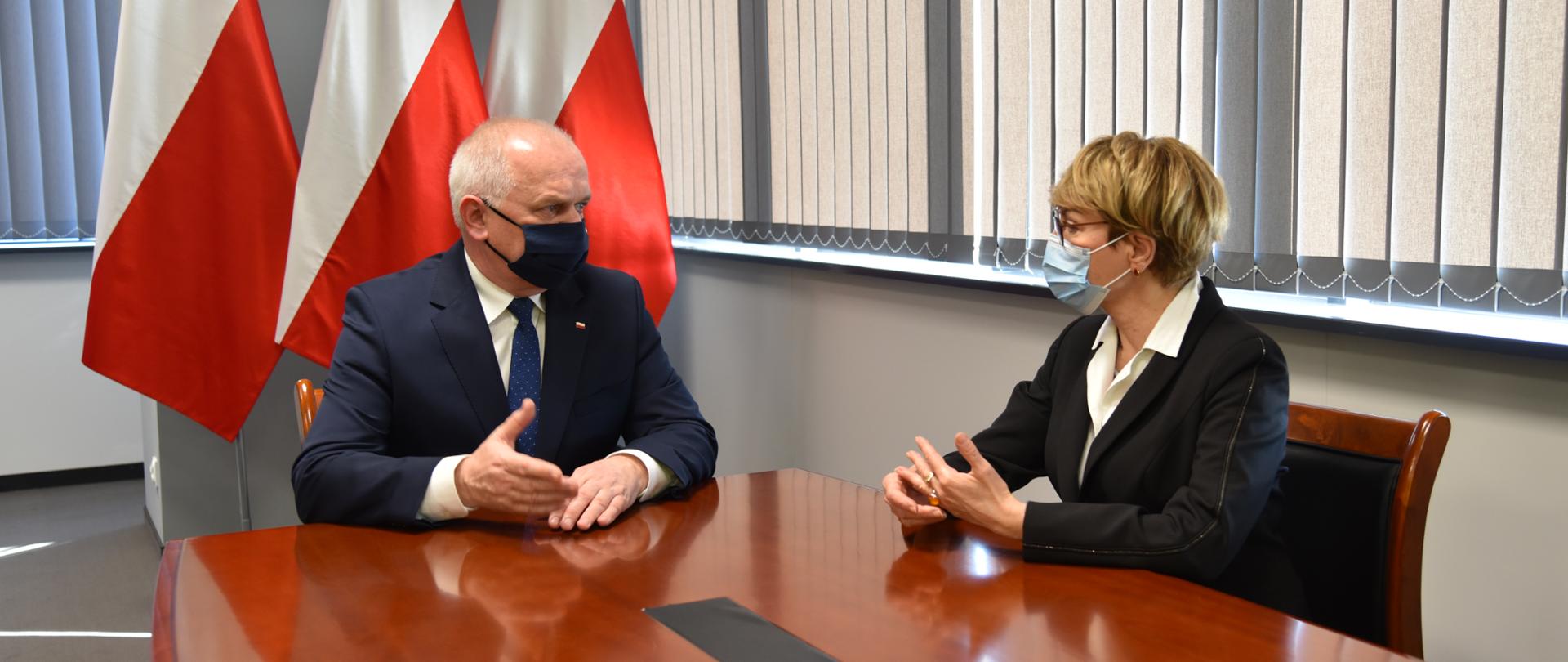 Wojewoda Władysław Dajczak rozmawia w swoim gabinecie z marszałek Elżbietą Polak. Oboje siedzą przy stole, w tle biało-czerwone flagi i okna zasłonięte szarymi żaluzjami 