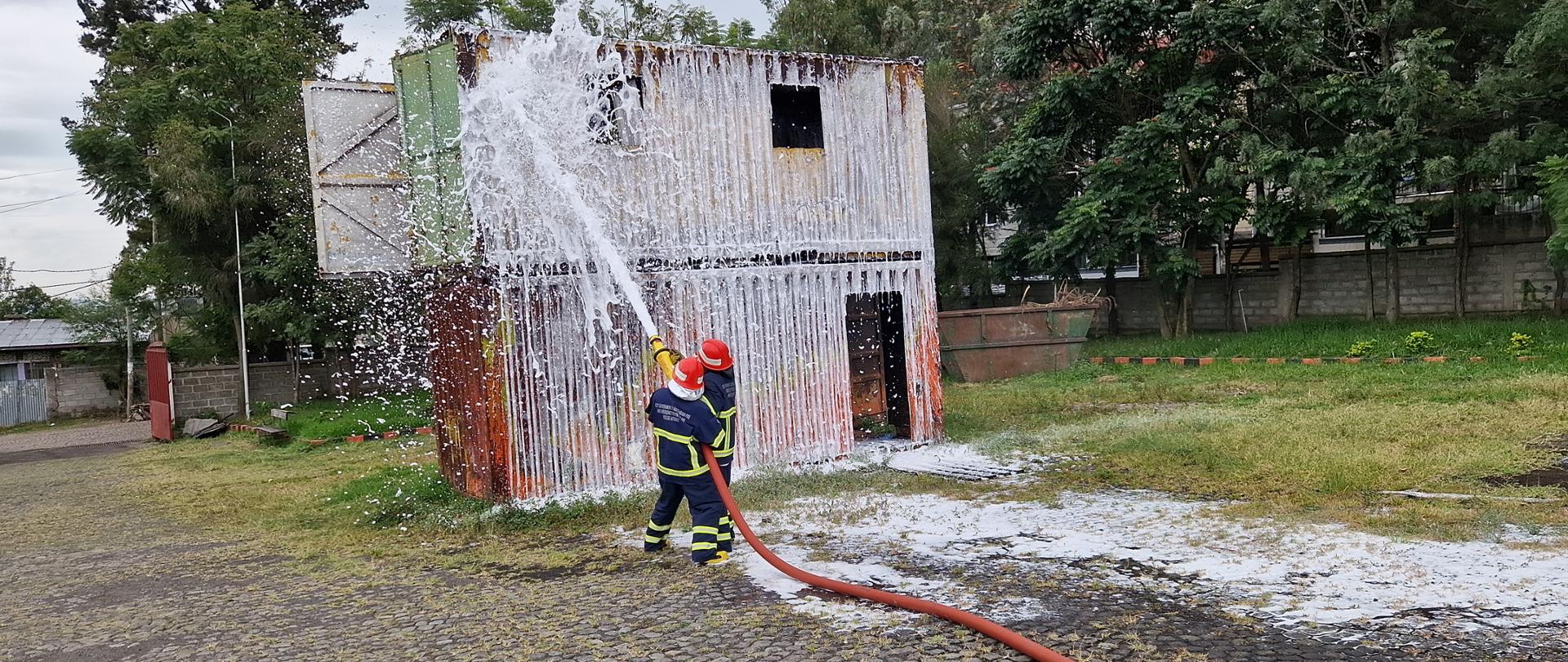 Dwie osoby w strojach strażackich oblewają wodą z węża metalowy kontener