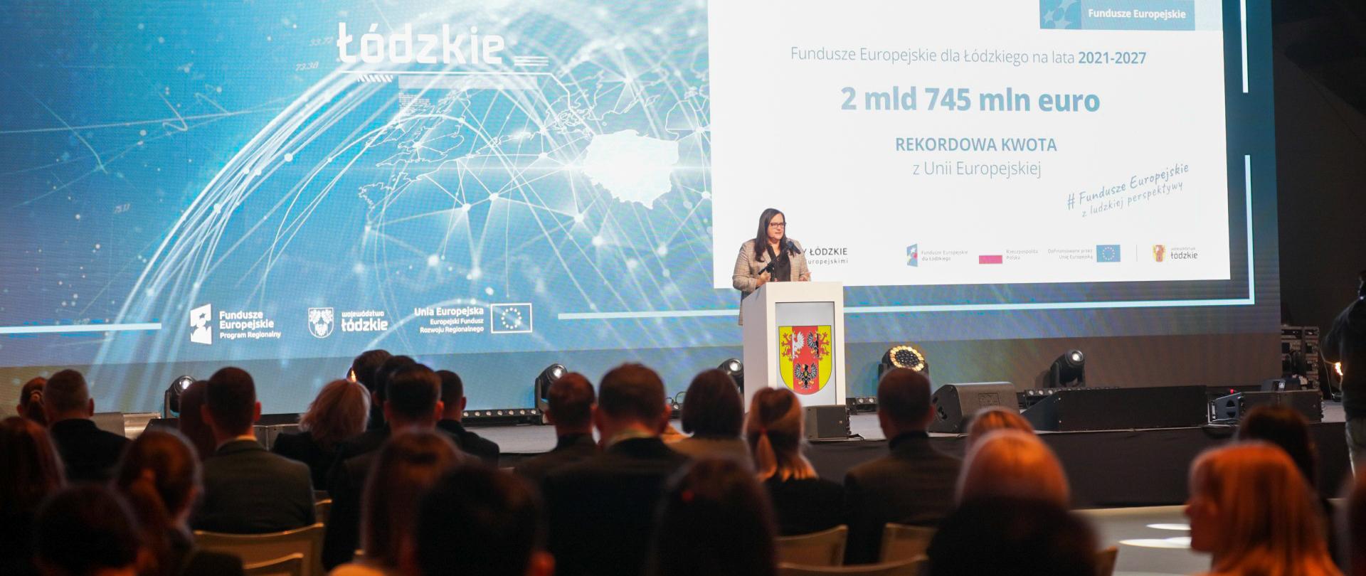 Wiceminister Małgorzata Jarosińska-Jedynak w sali konferencyjnej stoi w mównicy z mikrofonem. Przed nią publiczność. Za nią na ekranie napis "2 mld 745 mln euro".