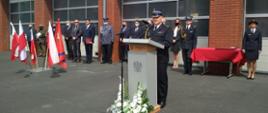 Na zdjęciu strażak PSP w mundurze wyjściowym przemawia stoi za mównicą, w tle zaproszeni goście oraz bramy garażowe strażnicy.
