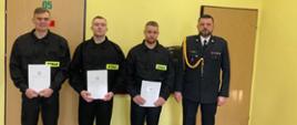Na tle żółtej ściany stoi trzech nowych strażaków ubranych w umundurowanie koszarowe oraz zastępca komendanta w mundurze wyjściowym.