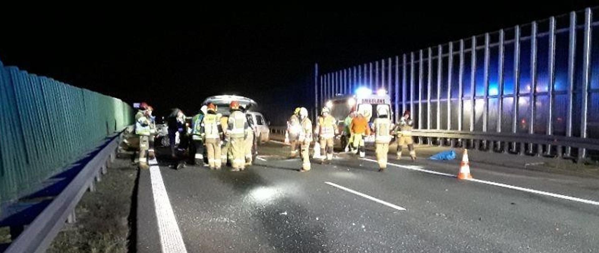 Na zdjęciu samochody znajdujące się na autostradzie po wypadku. Przed nimi strażacy w ubraniach specjalnych prowadzący czynności.