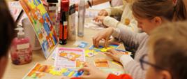 Przy stole grupa dzieci układa kolorowe puzzle o tematyce bezpieczeństwa kosmetyków. Na blacie widoczne są elementy puzzli, ilustracje z symbolami ostrzegawczymi oraz różne opakowania kosmetyków i pojemniki z dozownikami, wykorzystywane jako materiały edukacyjne.