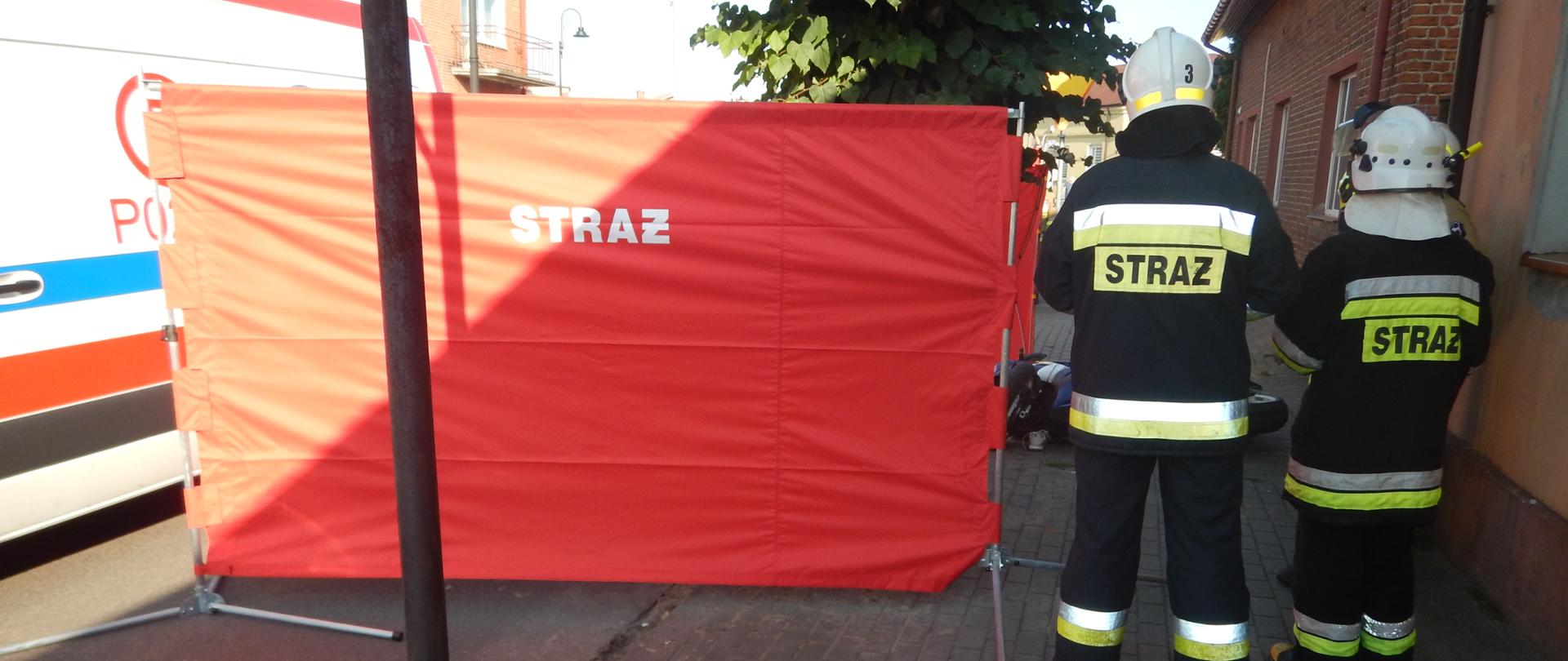 Zdjęcie przedstawia strażaków stojących obok parawanu zasłaniającego miejsce zdarzenia.