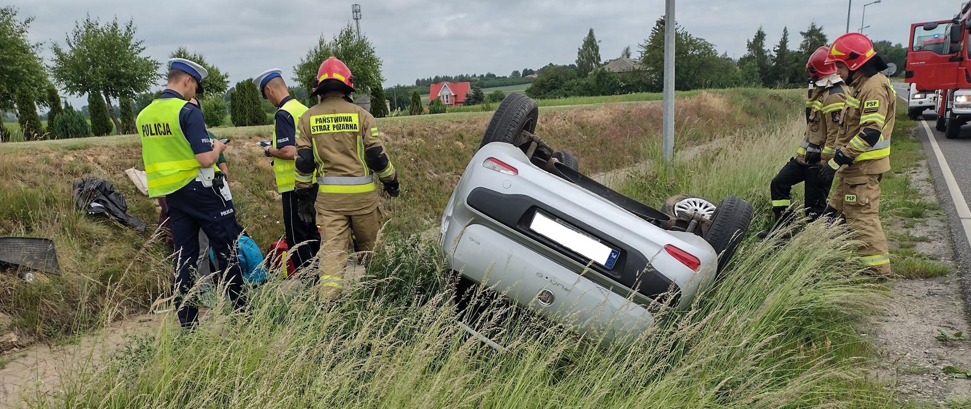 Dachowanie samochodu osobowego na drodze wojewódzkiej nr 751 w miejscowości Szwarszowice w gm. Bodzechów. Na zdjęciu widać samochód leżący w rowie przy drodze, który dachował. Wokół samochodu czynności ratownicze prowadzą strażacy oraz policjanci. W tle widać zaparkowane pojazdy gaśnicze. Zdjęcie wykonane w porze dziennej. Niebo pochmurne.