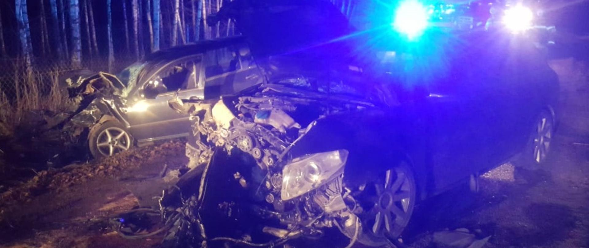 Dwa rozbite pojazdy osobowe. Jeden z pojazdów znajduję się w rowie i ma rozbity cały przód i wybitą szybę boczną od strony kierowcy, 2 pojazd stoi na drodze i ma też rozbity przód . Za nimi stoi na sygnałach samochód gaśniczy. 