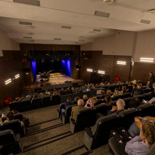 scena w domu kultury w głubczycach, zdjęcie wykonane z punktu bardzo oddalonego od sceny, perspektywa widza siedzącego na tyle sali, widoczne osoby siedzące na publiczności