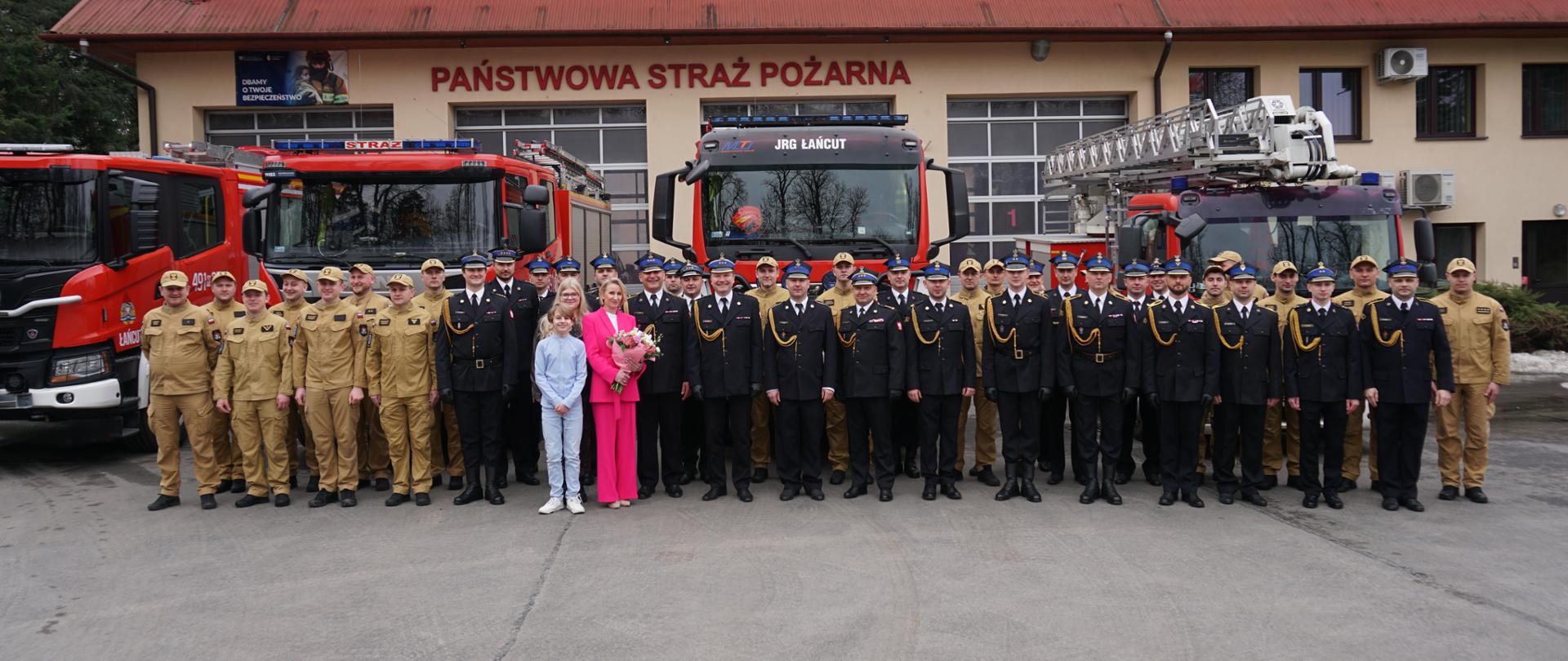 Na zdjęciu widoczni są wszyscy uczestniczący w uroczystości przejścia na zaopatrzenie emerytalne strażaków na tle samochodów ratowniczo-gaśniczych. W tle znajduje się Komenda Powiatowa PSP w Łańcucie.