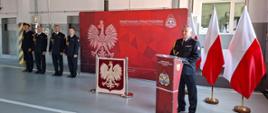 Na garażu obok flag Polski i banera Państwowej Straży Pożarnej stoją w rzędzie strażacy ubrani w mundury galowe. Przed nimi wystawione jest Godło Polski. Za mównicą stoi Komendant również ubrany w mundur galowy.