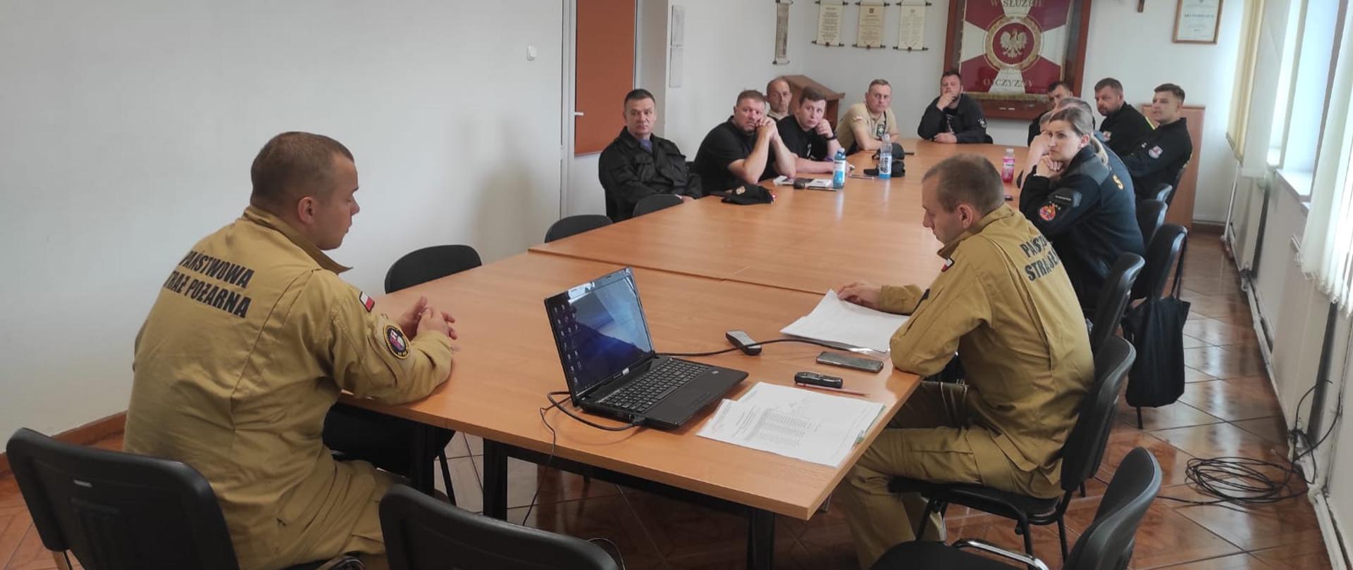 Zdjęcie przedstawia wykład szkolenia dowódców OSP strażacy w piaskowych mundurach siedzą przy stołach z dokumentami i papierami opowiadając druhom osp