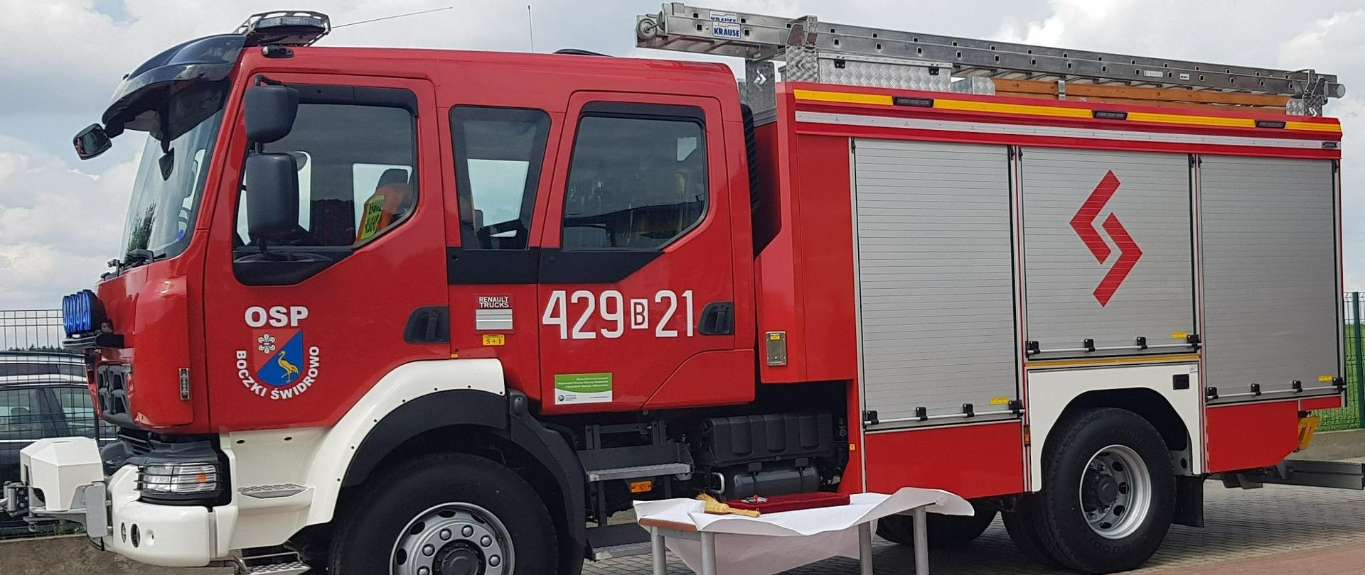 Nowy samochód gaśniczy OSP stoi na placu. Przed nim stolik z białym nakryciem.